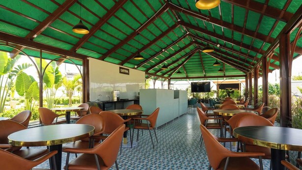 A covered outdoor seating area with tables and chairs, a kitchen area, and a view of greenery. - Palette Haritha Wellness Resort CheckIn (Sangareddy)