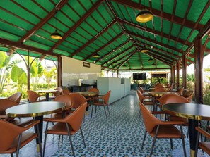 A covered outdoor seating area with tables and chairs, a kitchen area, and a view of greenery.