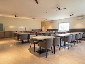 Un comedor amplio con varias mesas y sillas, ventiladores de techo y un ventanal que proporciona luz natural.