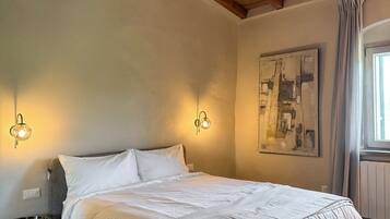 A bedroom with a large bed, wooden ceiling beams, wall-mounted lighting, a painting, and a window with curtains.