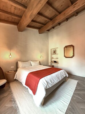 A bedroom with a large bed, wooden ceiling beams, a bedside table, a chair, and a wall-mounted mirror. - Vrbo Property (Riparbella)
