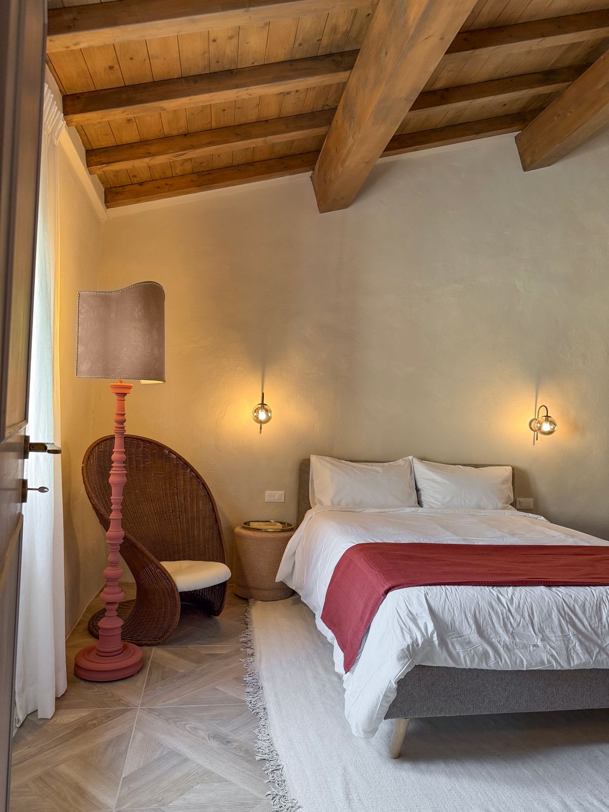 A bedroom with a wooden ceiling, a bed with white bedding, a red throw, a wicker chair, a floor lamp, and two wall sconces.
