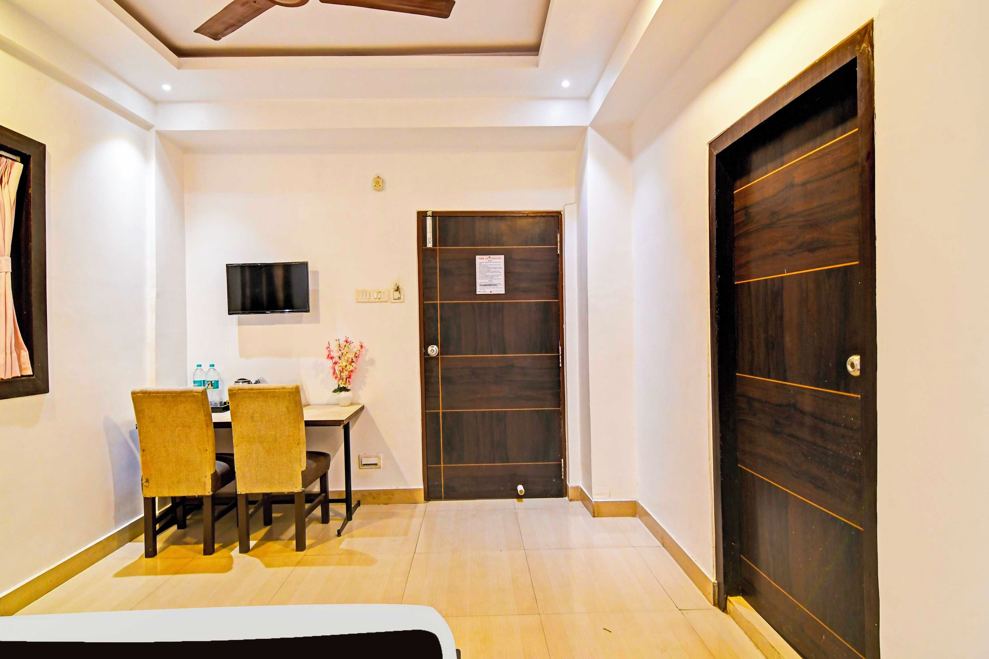 Habitación de hotel con cama, escritorio con televisor, puerta y una mesita con un jarrón de flores.