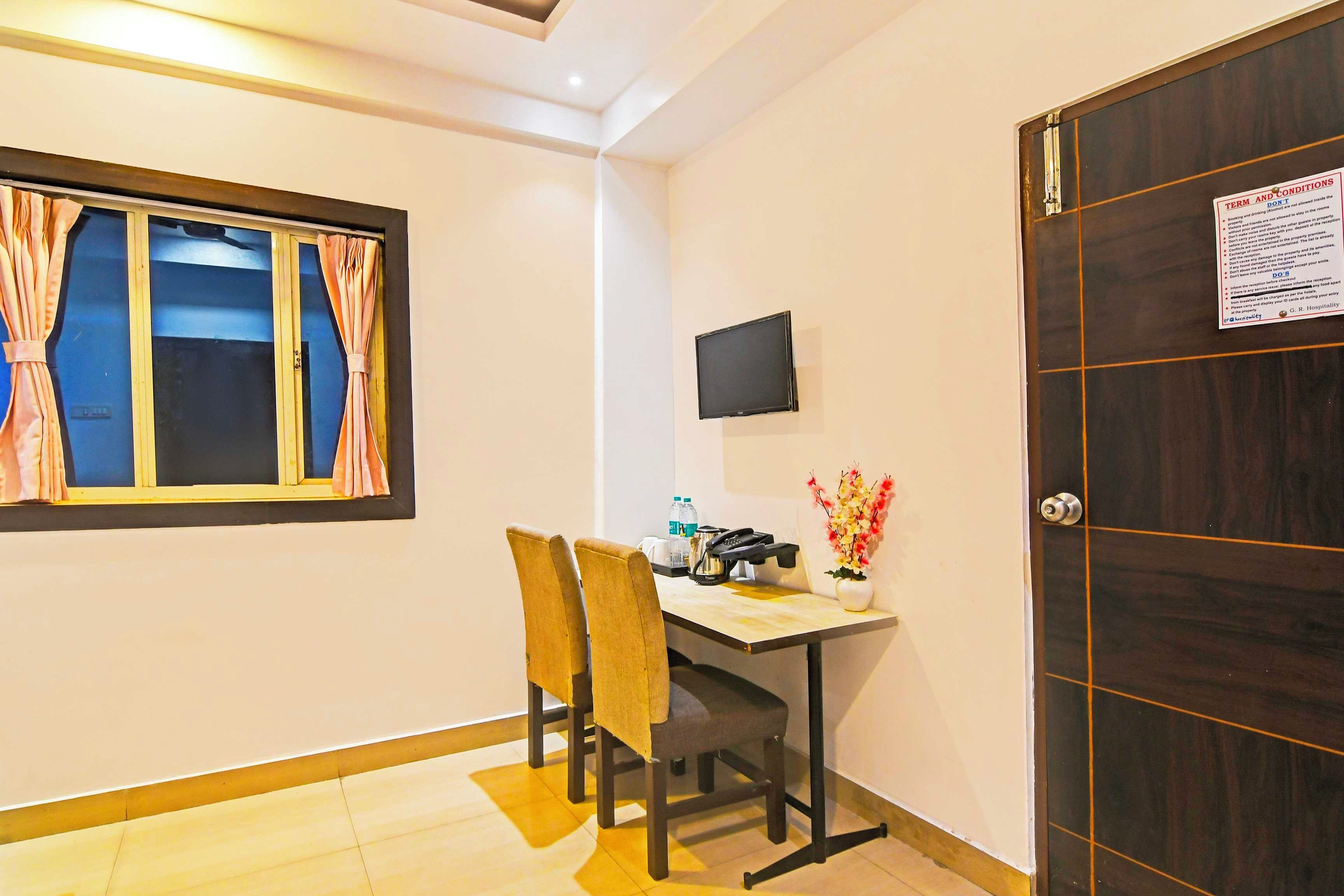 Habitación de hotel con escritorio, sillas, televisor de pantalla plana, puerta de madera oscura y ventana con cortinas.