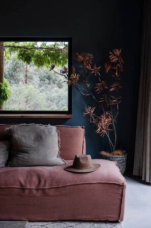 A room with a dark blue wall, a window with a view of greenery, a pink sofa with cushions, and a decorative plant.