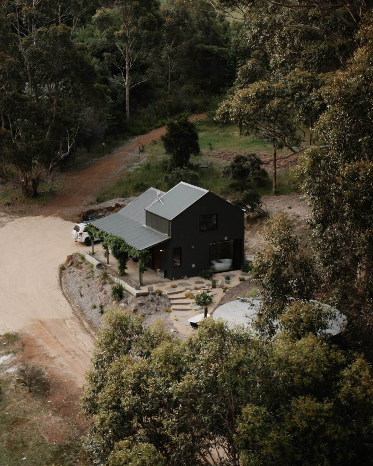 A two-story house with a sloped roof, surrounded by dense vegetation and a dirt driveway.