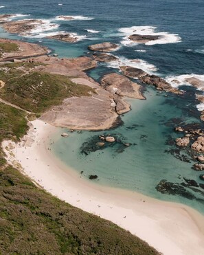 An aerial view of a coastal area with a sandy beach, rocky outcrops, and clear blue waters.