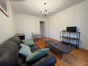 Een woonkamer met een bank, een salontafel, een eettafel met stoelen, een televisie op een plank en een kroonluchter.