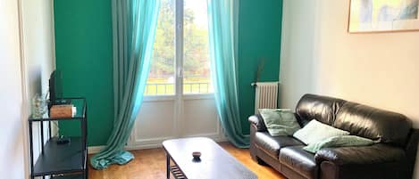 A room with a wooden floor, a black leather sofa, a coffee table, a black TV stand, and a window with curtains.