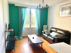 Een kamer met een houten vloer, een zwarte leren bank, een salontafel, een zwarte tv-meubel en een raam met gordijnen.