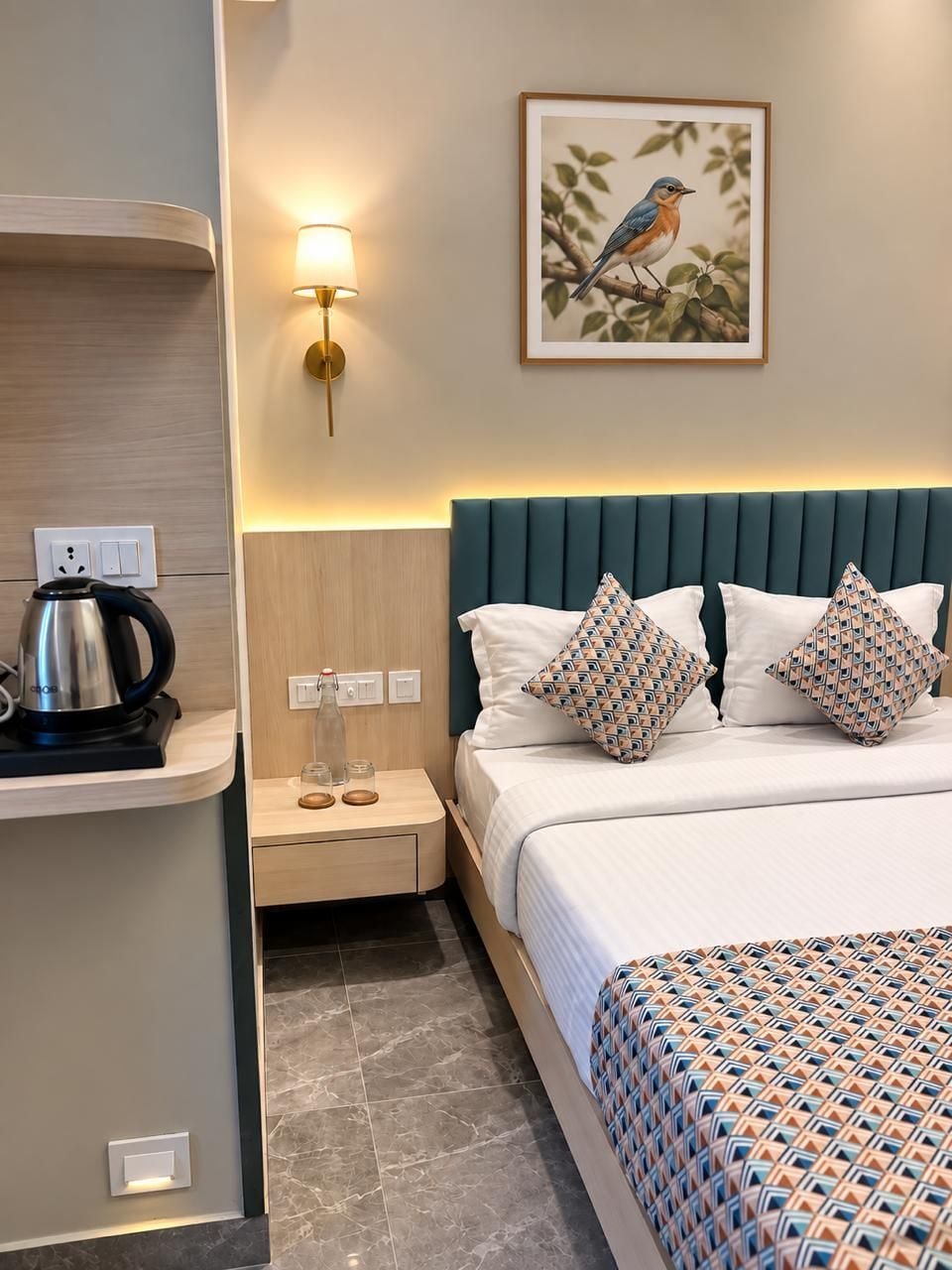 A hotel room with a bed, a nightstand, a kettle, and a wall-mounted lamp.