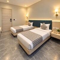 Habitación de hotel con dos camas, una puerta de madera, una mesita de noche con lámpara, y una lámpara fijada en la pared.