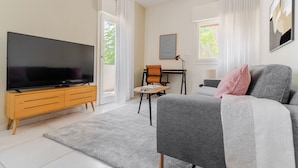 Una sala moderna con un televisor de pantalla plana sobre un mueble de madera, un sofá gris, una mesita con una silla y un ventanal con cortinas.
