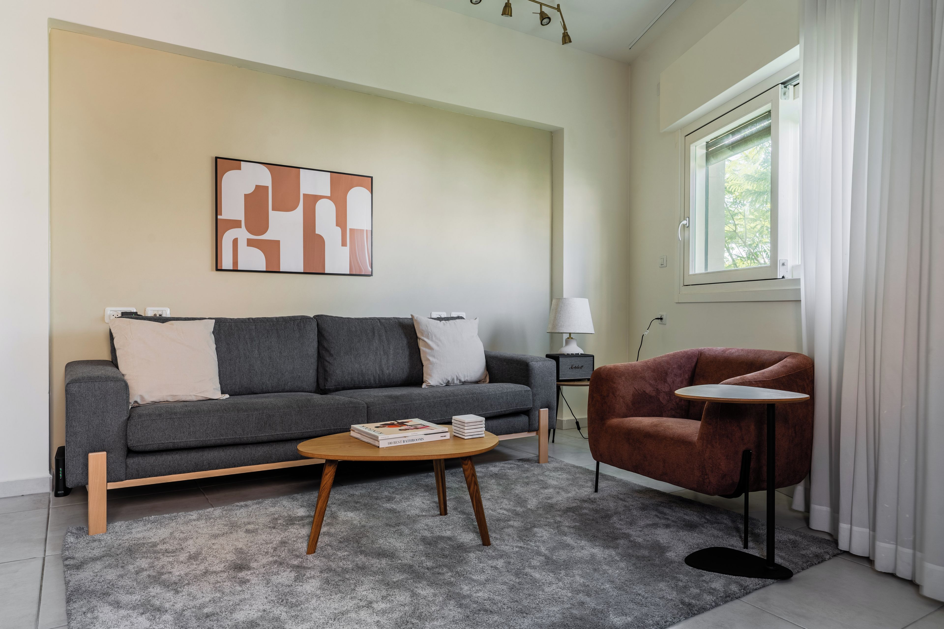 Una sala de estar moderna con un sofá gris, una mesa redonda y un sillón rojo. Hay una lámpara en una mesita auxiliar y una estantería llena de libros.