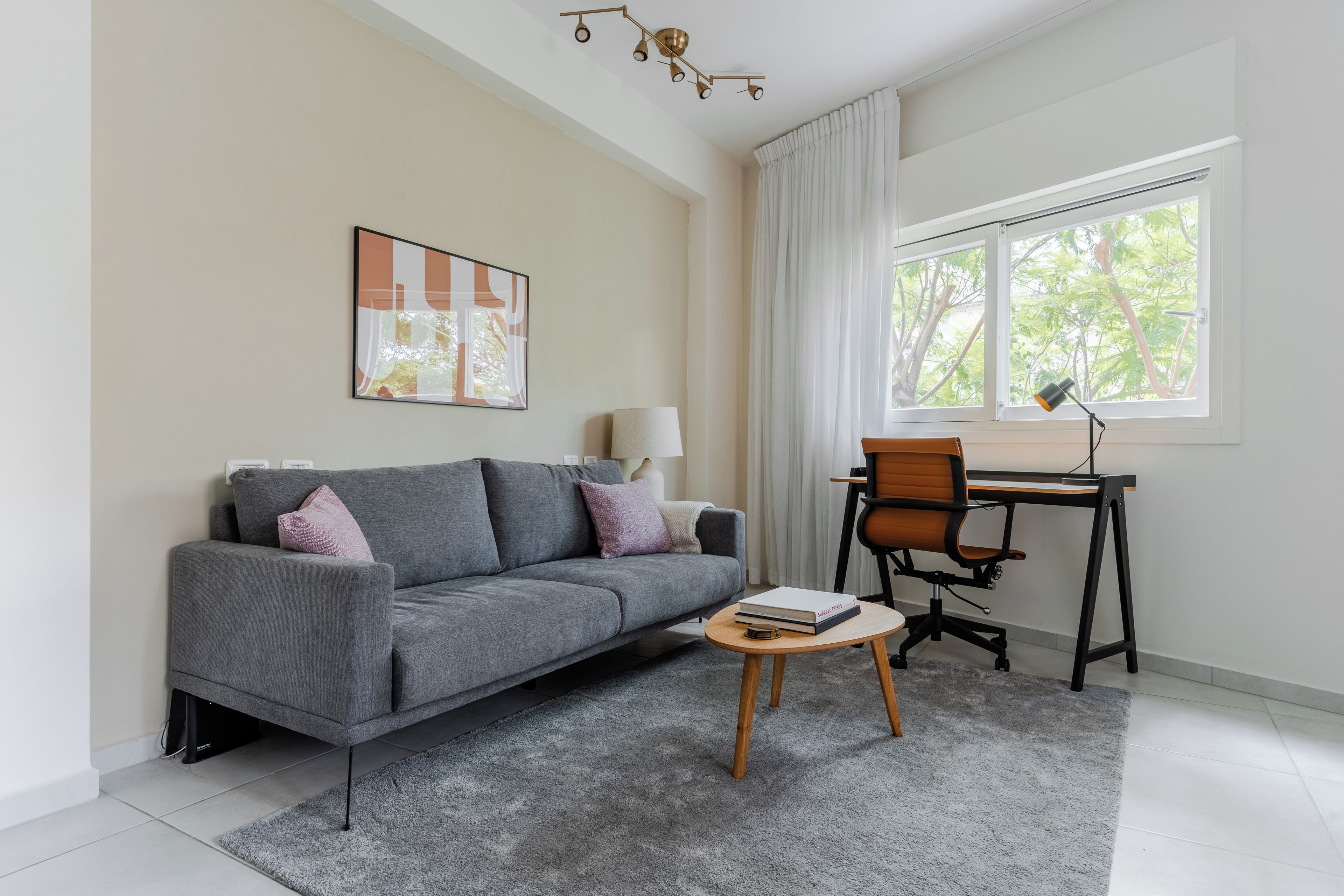 Una sala de estar moderna con un sofá gris, una mesita de centro redonda, un escritorio con una silla y un ventanal con cortinas.