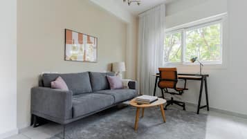 A modern living room with a grey sofa, a small round coffee table, a desk with a chair, and a large window with curtains.