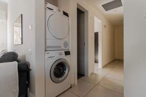 A laundry room with a front-loading washing machine and dryer, a small seating area with a chair and a basket, and a hallway leading to other rooms. - Blueground Nahalat Bynamin City view (Tel Aviv-Yafo)