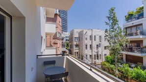 Un balcón con mesa y sillas, con vista a un paisaje urbano de varios edificios y cielo despejado.