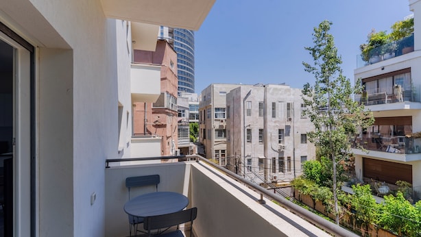 Un balcón con mesa y sillas, con vista a un paisaje urbano de varios edificios y cielo despejado.