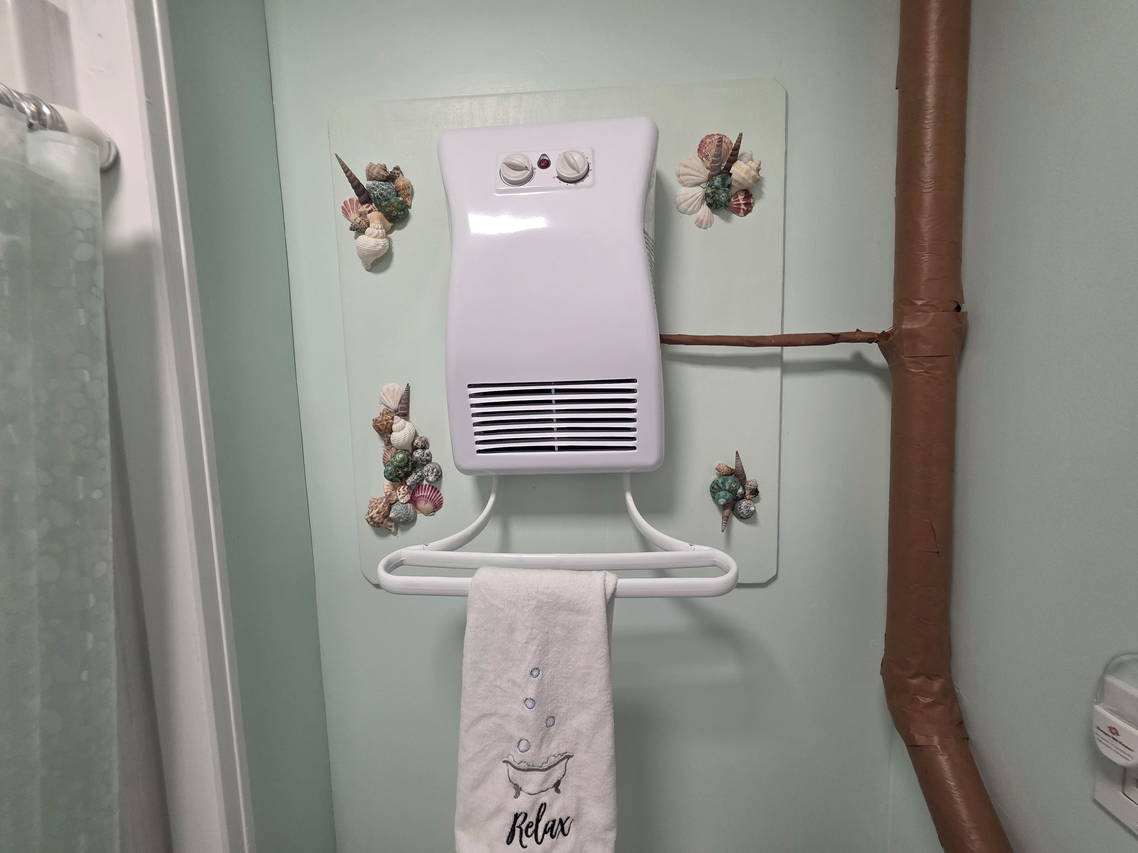 Une salle de bain avec un porte-serviettes sur lequel est posée une serviette ornée du mot «Relax», un radiateur blanc fixé au mur et des coquillages décoratifs.