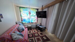 Quarto com cama, televisão, mesinha e janela com cortinas.