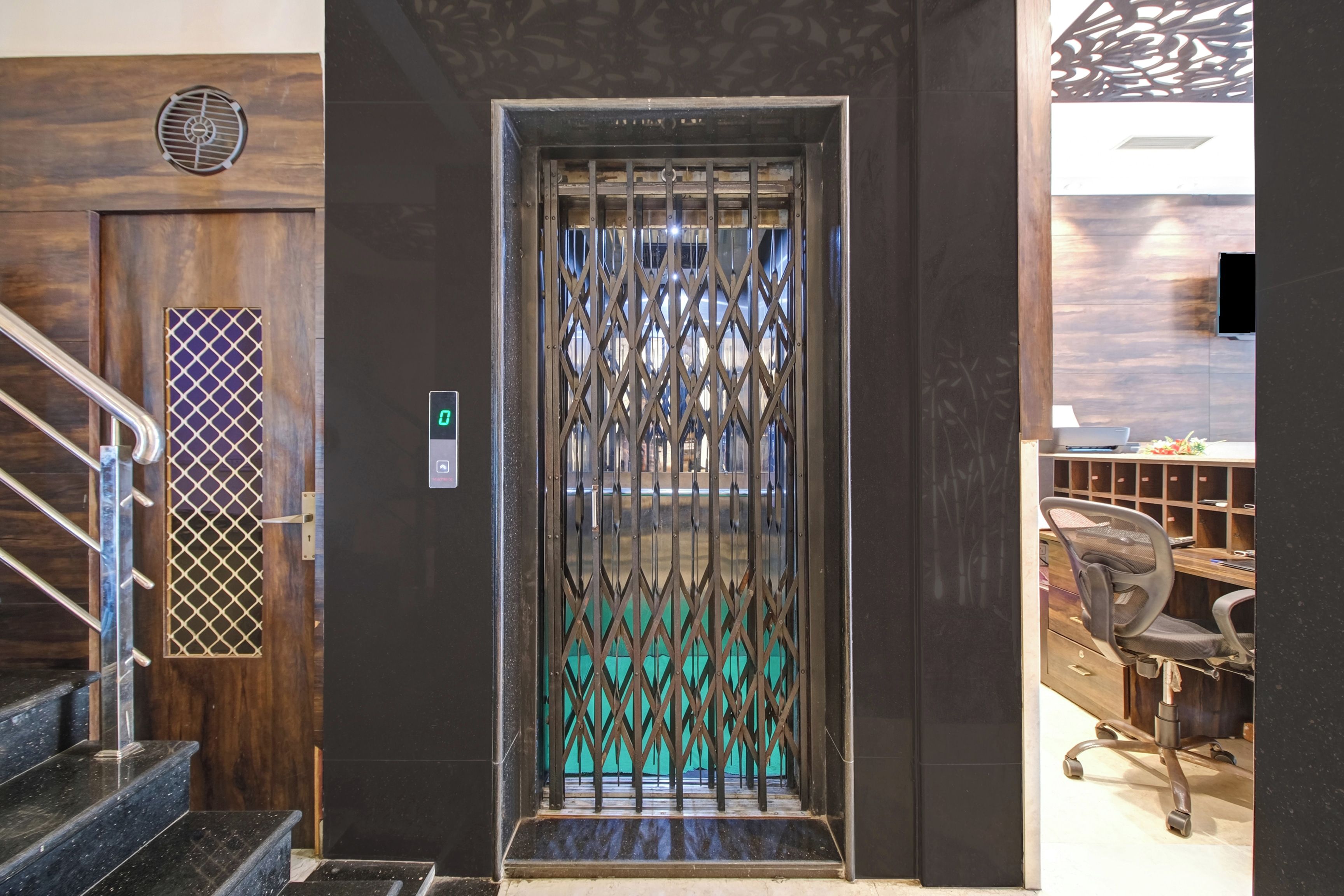 En modern hiss med en dörr av metallgaller, en väggpanel av trä med en ventilationsöppning och en trappa med räcken av metall.