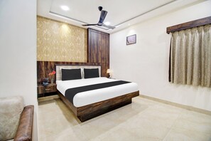 Quarto de hotel com cama, mesa de cabeceira, escrivaninha, cadeira, ventoinha no teto, janela com cortinas e um quadro na parede.