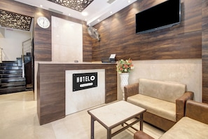 Área de recepção de hotel com balcão, sofá, mesa de centro, relógio e TV fixada na parede.