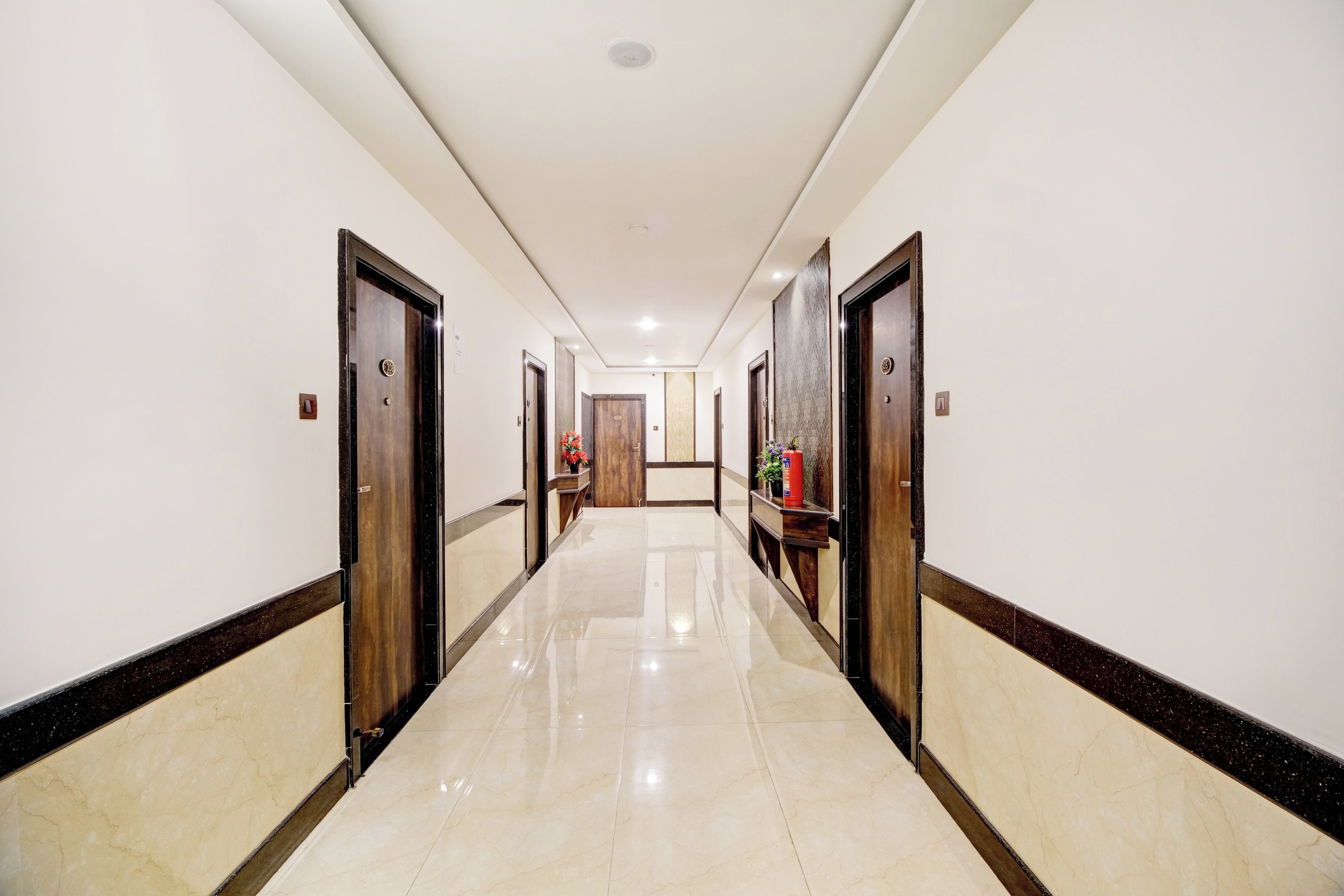 Corredor de hotel com piso de mármore polido, portas de madeira e um extintor de incêndio na parede.