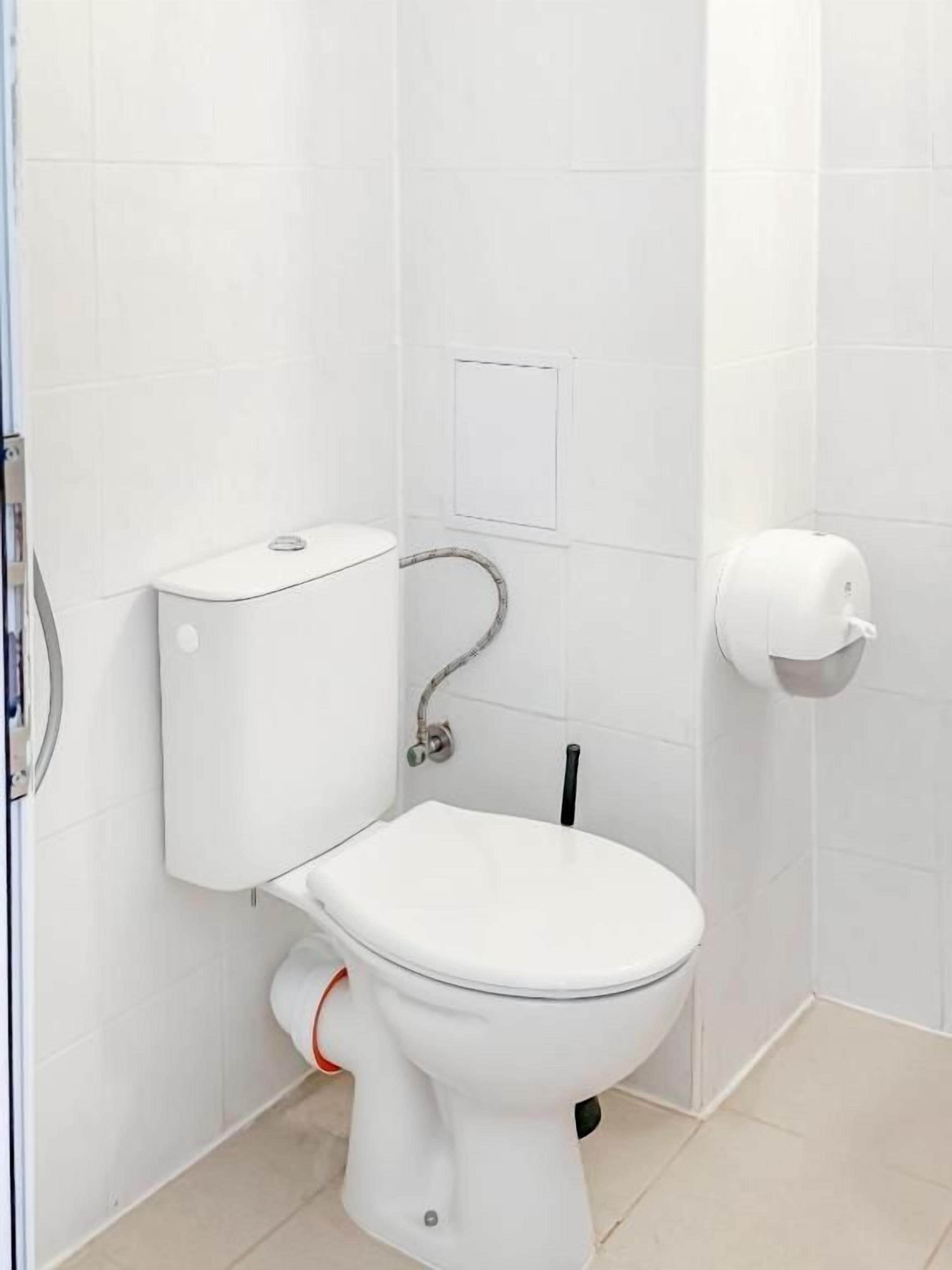 Inodoro blanco estándar con manija de descarga, un dispensador de papel higiénico y una tubería visible.