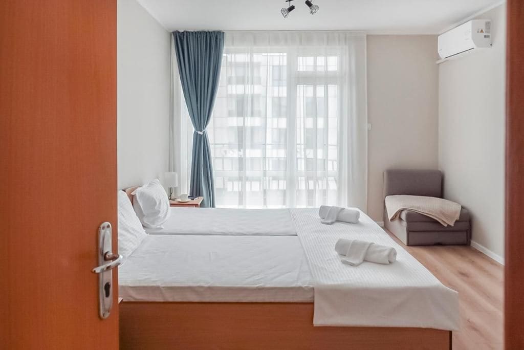 A hotel room with two beds, a window with curtains, a chair, and an air conditioning unit.