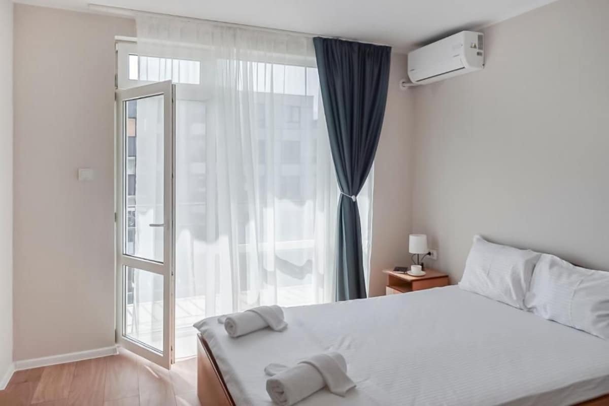 A hotel room with a bed, a nightstand, a lamp, an air conditioning unit, and a view through a window with curtains.