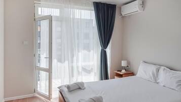 Habitación de hotel con cama, mesita de noche, lámpara, equipo de aire acondicionado y vistas a través de una ventana con cortinas.
