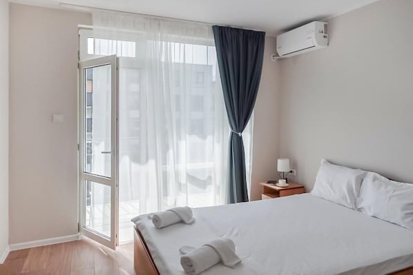 A hotel room with a bed, a nightstand, a lamp, an air conditioning unit, and a view through a window with curtains. - Solea Family Hotel (Ravda)