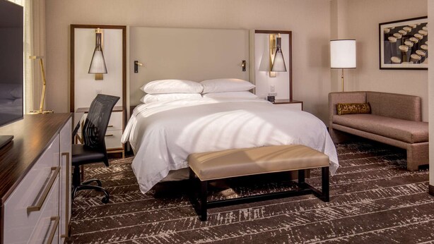 A hotel room with a bed, bedside table, lamp, chair, and a sofa. - Steps From Downtown + On-Site Dining, King Bed (Youngstown)