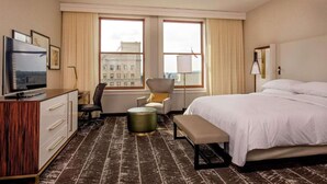 A hotel room with a bed, a desk with a TV, a chair, a lamp, and a view of buildings through the windows. - Steps From Downtown + On-Site Dining, King Bed (Youngstown)