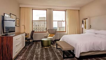 A hotel room with a bed, a desk with a TV, a chair, a lamp, and a view of buildings through the windows.