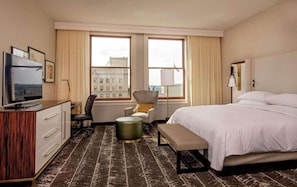 Habitación de hotel con cama, escritorio con televisor, silla, lámpara y vista a edificios a través de las ventanas.