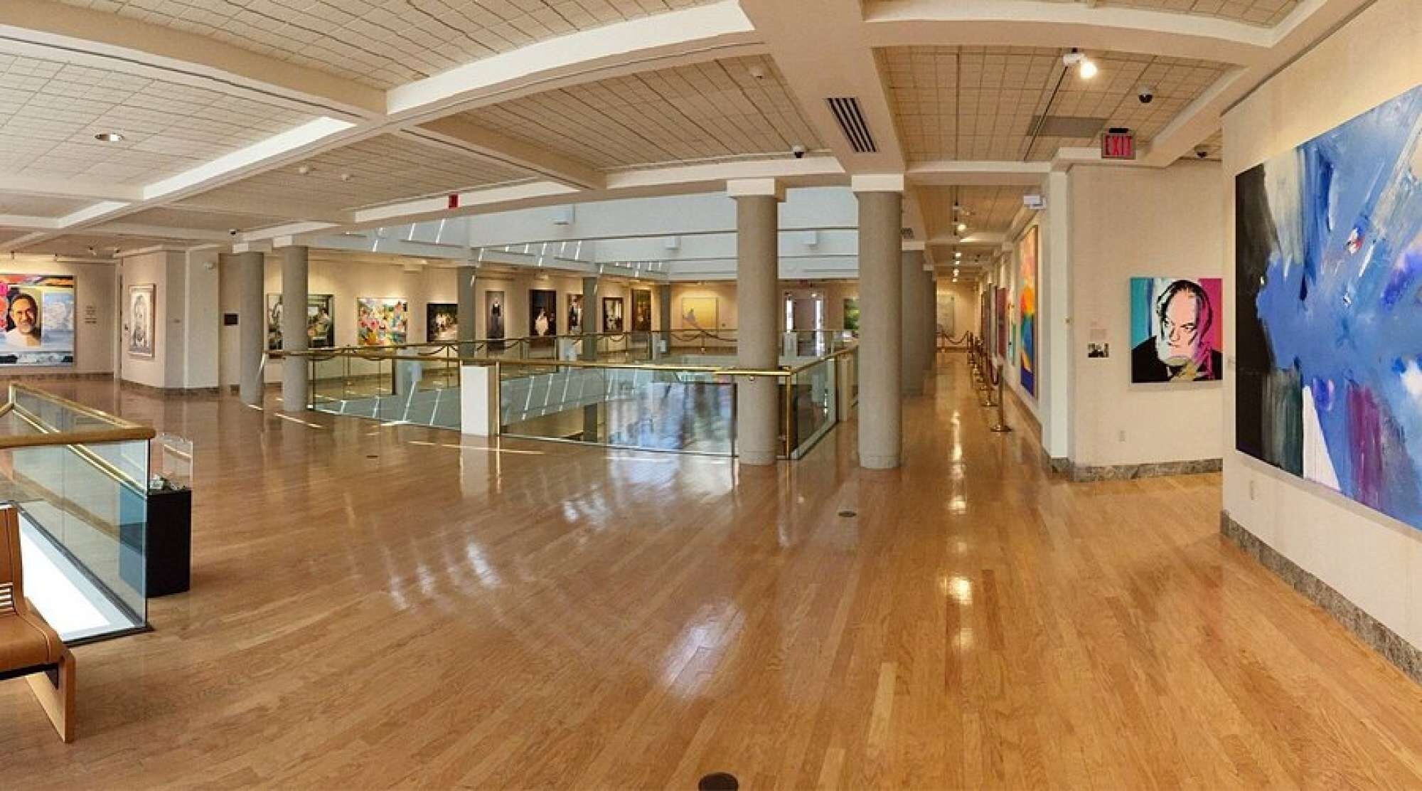 A spacious indoor area with a polished wooden floor, glass railings, and a series of artworks on the walls.