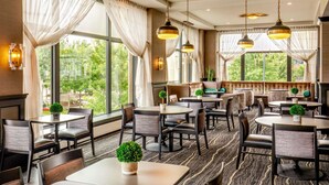 Zona pranzo di un hotel con diversi tavoli e sedie, ampie finestre con tende e lampadari pendenti.