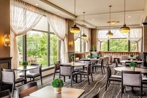 Comedor de hotel con varias mesas y sillas, ventanas amplias con cortinas, y lámparas colgantes.