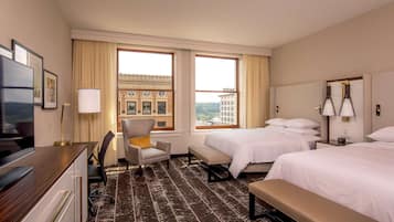 A hotel room with two beds, a desk with a chair, a bench, a TV, and a large window with a view of buildings.