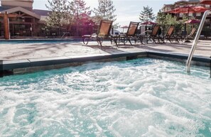Ein Whirlpool, umgeben von Liegestühlen und Sonnenschirmen, mit einem Pool und Gebäuden im Hintergrund.