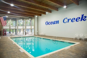 An indoor swimming pool with a view of the outdoors, a large American flag, and the name 'Ocean Creek' on the wall. - 1BR Villa Near Beach + On-site Dining, Pool & Gym (Myrtle Beach)