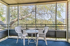 A covered outdoor seating area with a table and four chairs, overlooking a landscape with trees and a building in the background. - 1BR Villa Near Beach + On-site Dining, Pool & Gym (Myrtle Beach)