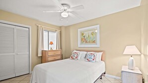 Quarto com cama, mesa de cabeceira, ventilador de teto, janela com cortinas, porta e um quadro pendurado na parede.