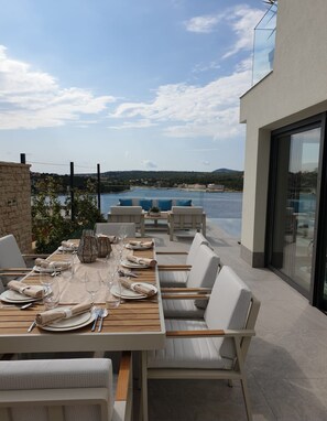 Un espace extérieur aménagé pour prendre les repas, offrant une vue sur la mer et les collines, avec des chaises blanches et une table en bois dressée pour un repas.