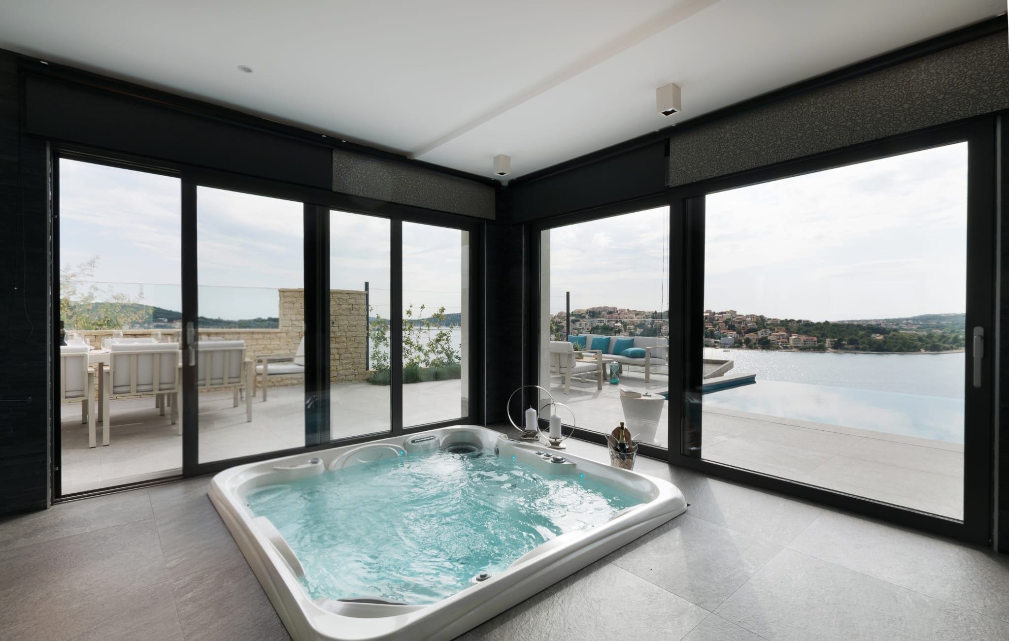 Une salle de bain moderne dotée d’une grande baignoire, offrant une vue sur la ville grâce à de larges fenêtres du sol au plafond, et d’un balcon aménagé avec du mobilier d’extérieur.