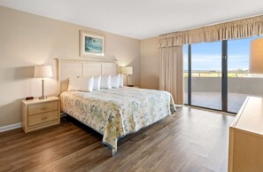 A hotel room with a bed, two bedside tables with lamps, a painting on the wall, and a balcony with a view. - Spacious 3BR Condo + Beach Access, Kitchen & Pools (Myrtle Beach)