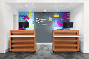 Zona d'admissió amb dues tauletes de recepció de fusta, una paret colorida amb dissenys abstractes i un cartell amb la inscripció 'Developer Inn MAINGATE'.